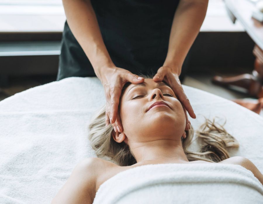 Blonde young woman on couch with white linen enjoys facial massage in spa